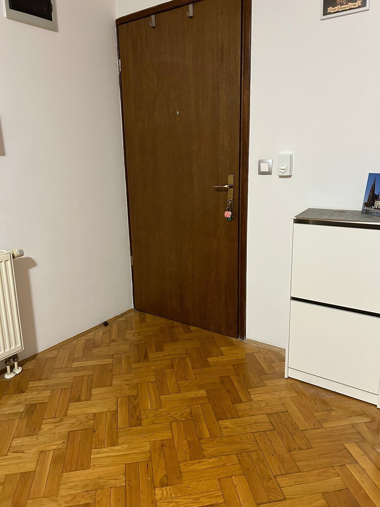 Une pièce avec une porte en bois, un radiateur et un meuble blanc sur lequel est posée une image.