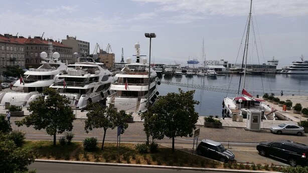 A marina with several yachts docked, a road in the foreground, and buildings in the background.