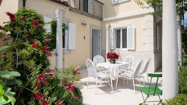 A courtyard with a white table and chairs, surrounded by greenery and a building with white shutters.