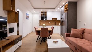 Un salon moderne avec un téléviseur, un canapé et une table basse. On y trouve également un coin repas avec une table et des chaises. La cuisine est visible et comprend des placards et des appareils électroménagers.