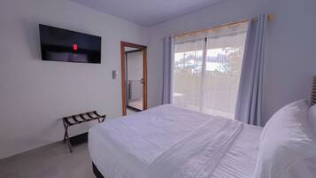 Una habitación de hotel con una cama grande, un banco, un televisor montado en la pared y un ventanal corredero que da a un balcón.