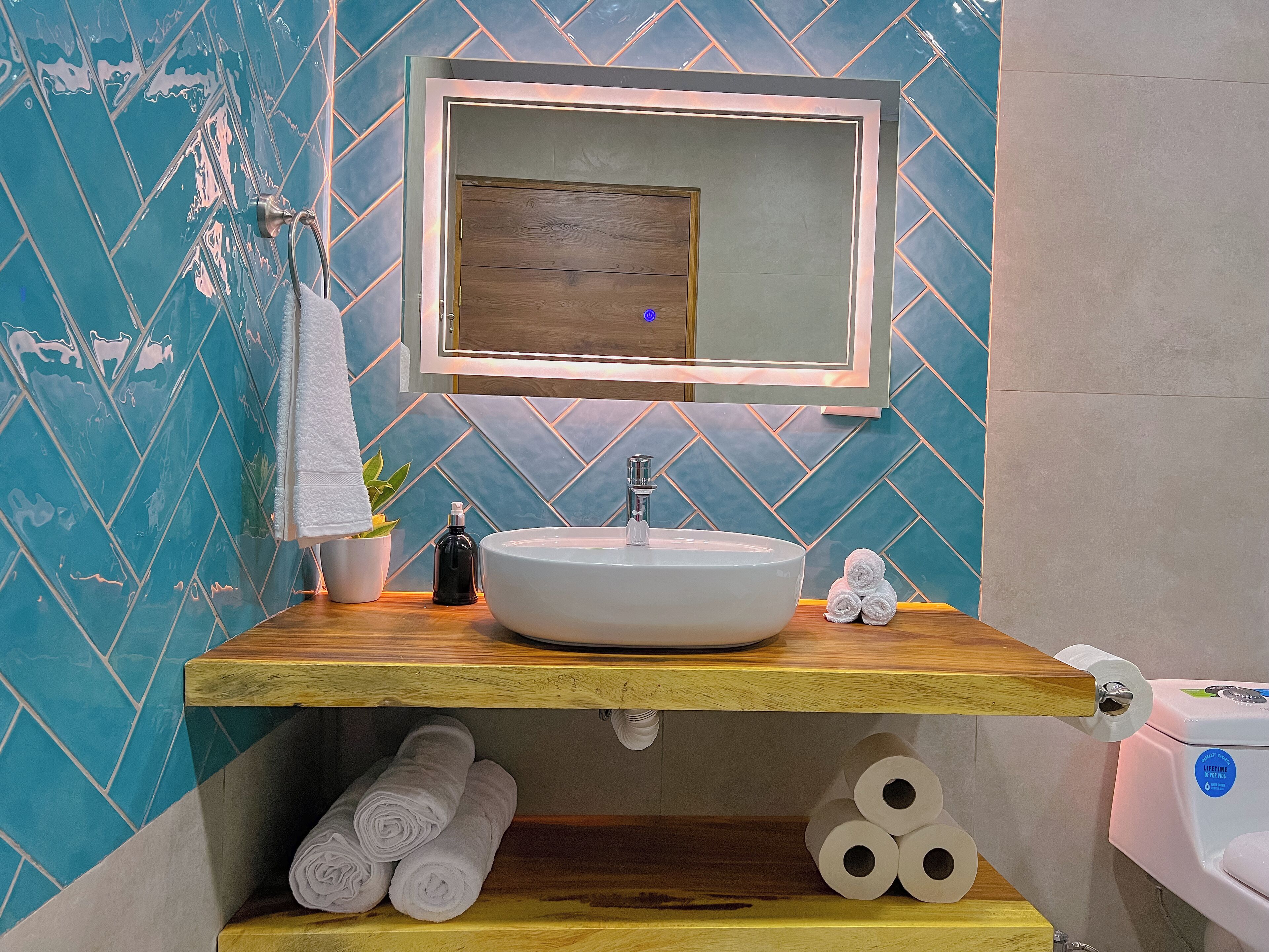 A bathroom with a wooden vanity, a white sink, and a mirror above it. There are towels and toilet paper on the vanity.