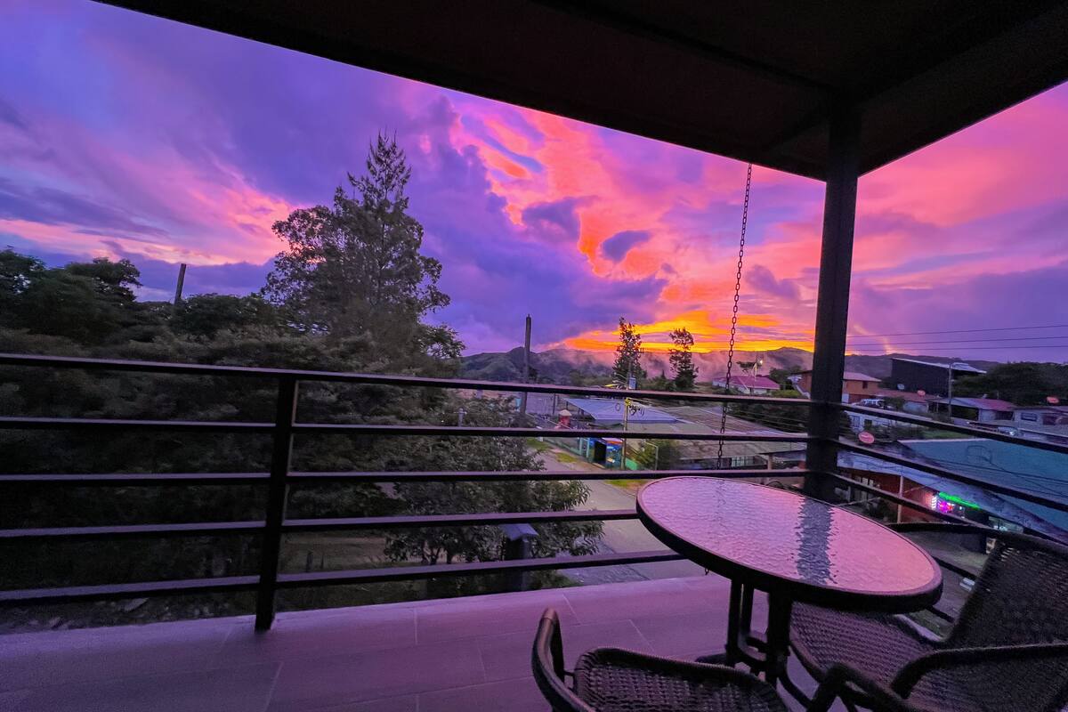 A balcony with a table and chairs, overlooking a scenic sunset view with mountains and trees.