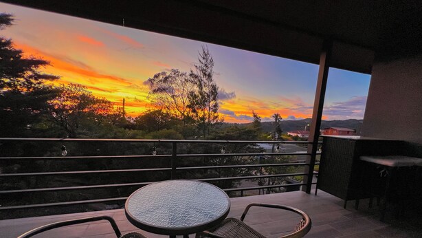 A balcony with a round table and two wicker chairs, overlooking a sunset with trees and a distant building. - Monstera Apartments Monteverde (Monteverde)