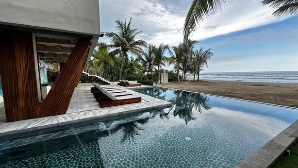 A modern infinity pool with wooden loungers, palm trees, and a beach view. - Charming Ocean line studio with largest pool in beautiful Troncones (Troncones)