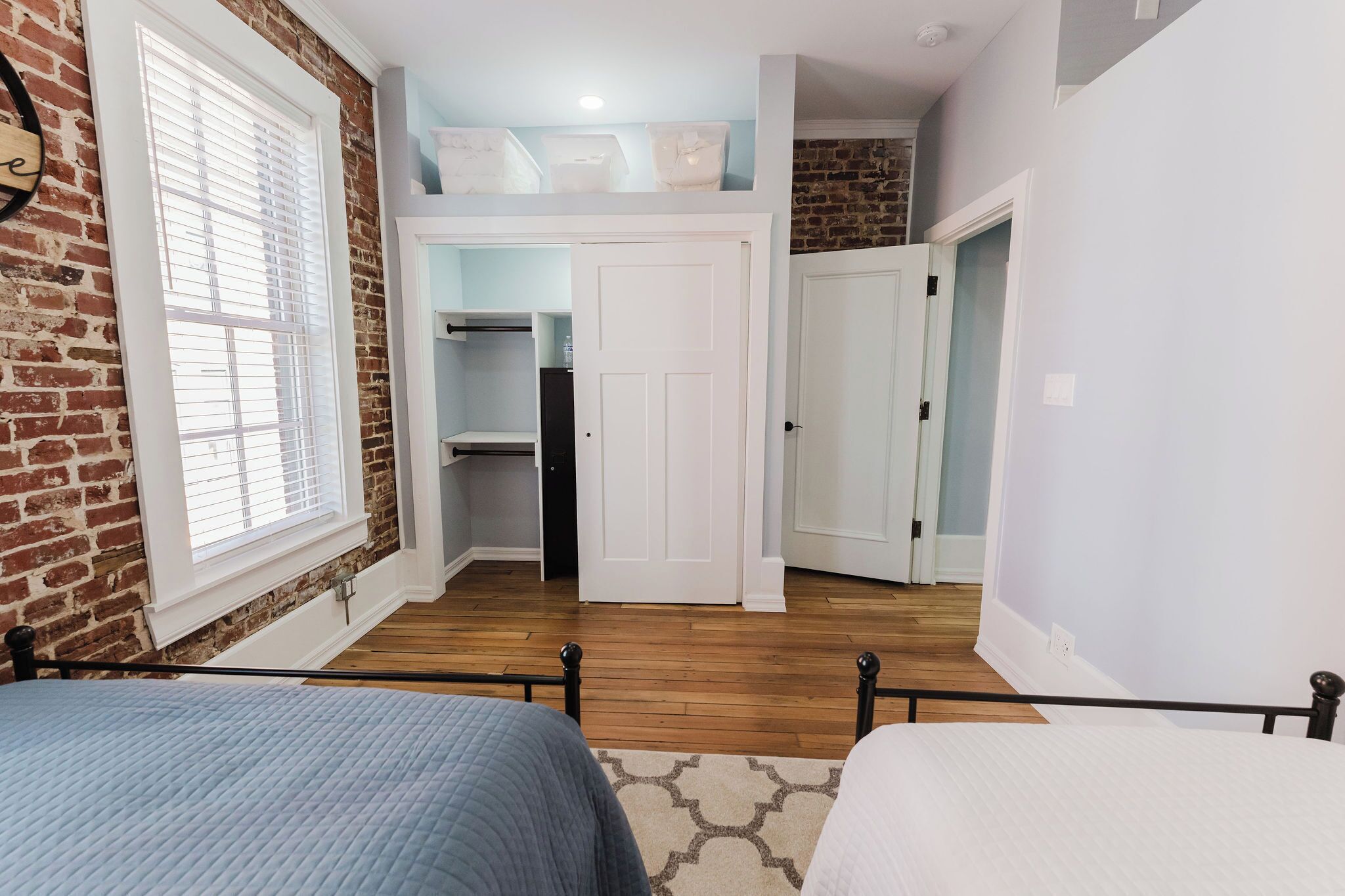 A bedroom with a brick wall, a window with blinds, a bed with a blue blanket, and a white door leading to a closet.