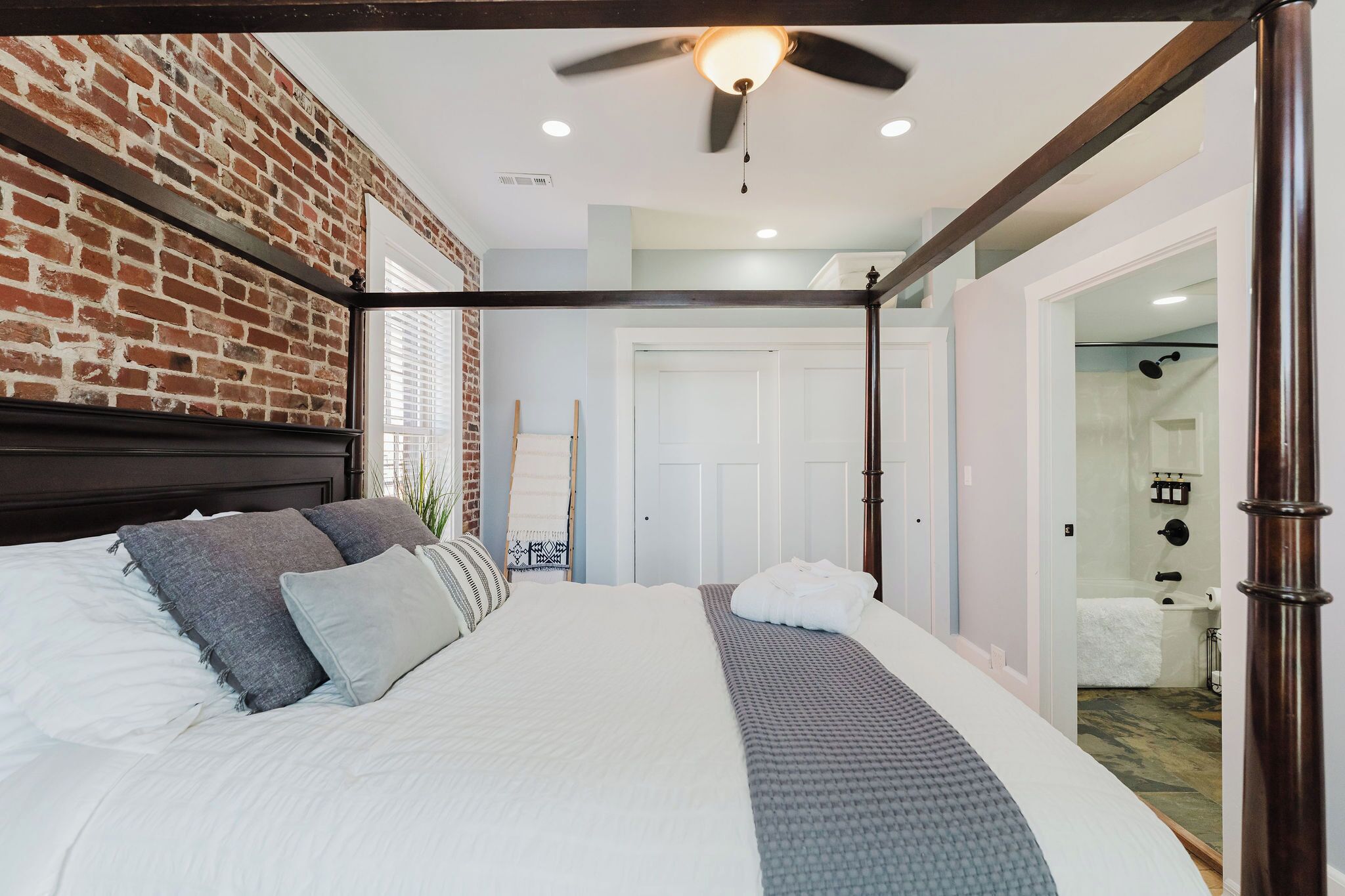 A bedroom with a brick wall, a ceiling fan, a bed with white bedding, and a bathroom visible through an open door.