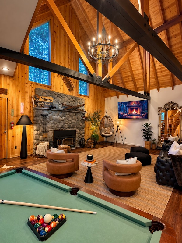 A cozy cabin interior with a pool table, comfortable seating, a stone fireplace, and a large window offering a view of the forest.