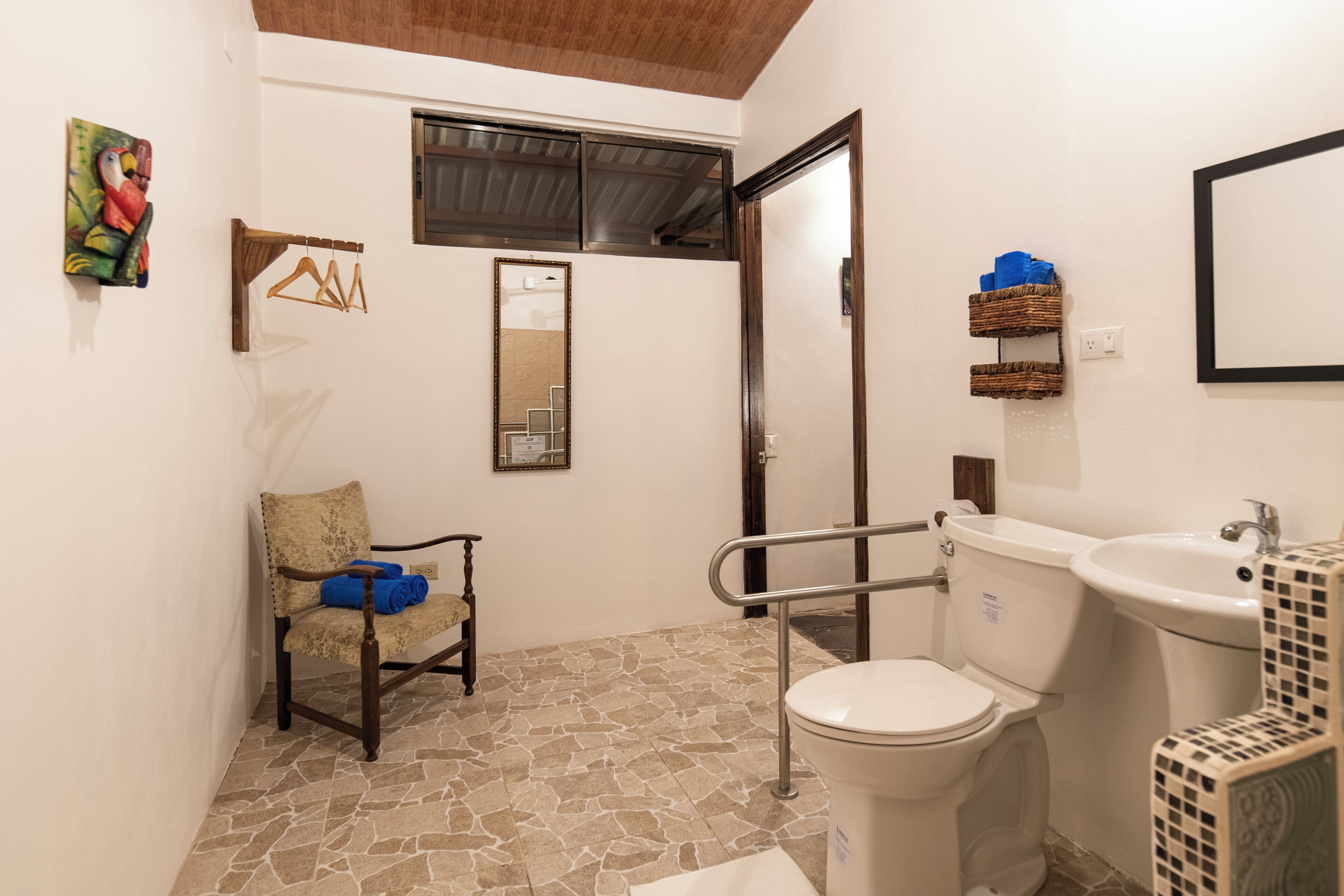 Ein Badezimmer mit Toilette, Waschbecken und Stuhl. Es gibt einen Spiegel, ein Fenster und ein Bild an der Wand.