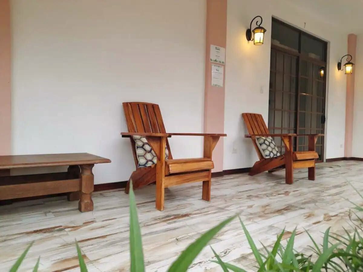 Un banco de madera y dos sillas de madera con cojines sobre un suelo de baldosas, con una lámpara de pared y una puerta al fondo.