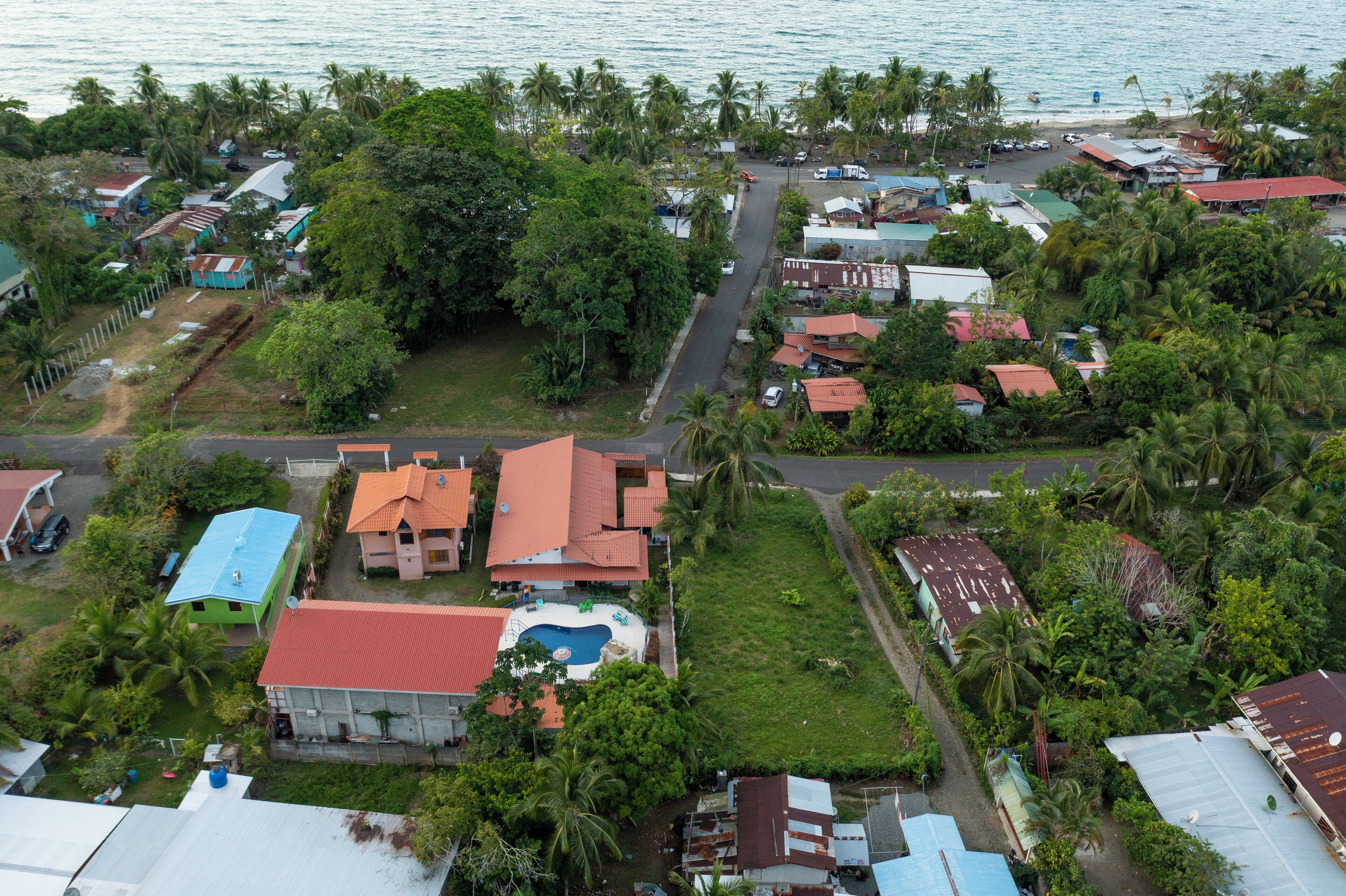 An aerial view of a coastal residential area with houses, a swimming pool, and palm trees.