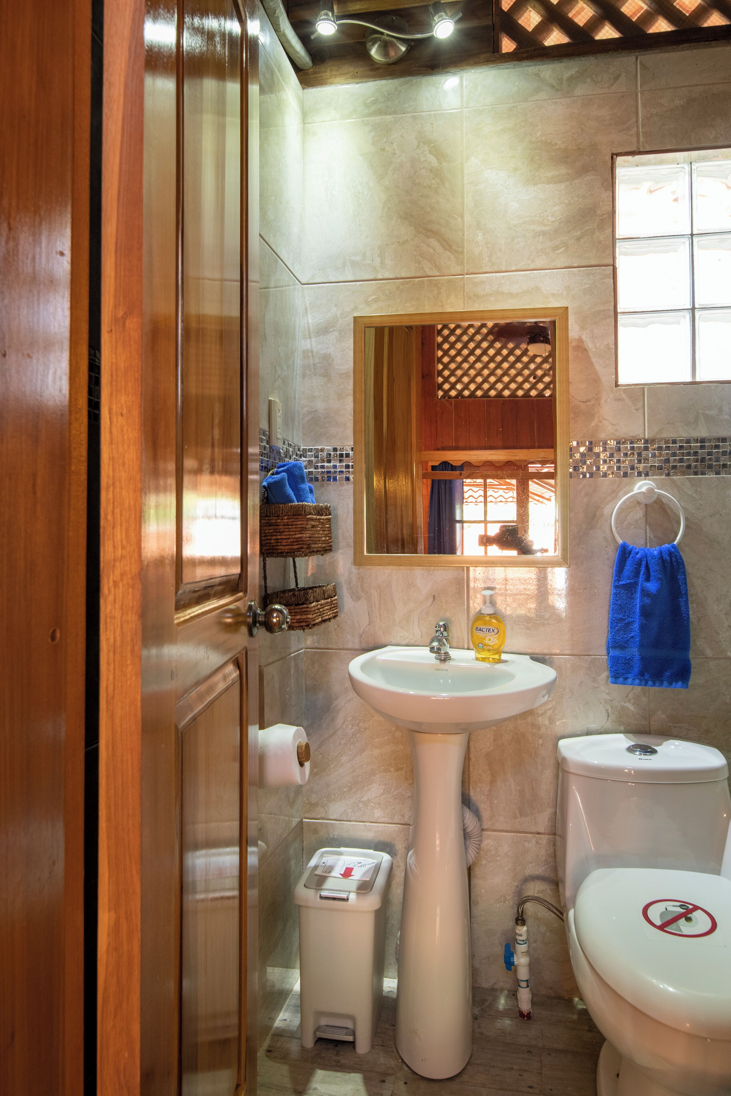 Un baño con lavamanos, inodoro y espejo. Se observa un perchero con una toalla azul.