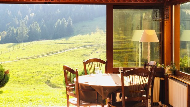 Un comedor de madera con una mesa y sillas, ventanas amplias que dan a un campo verde y árboles, y una lámpara en el alféizar.