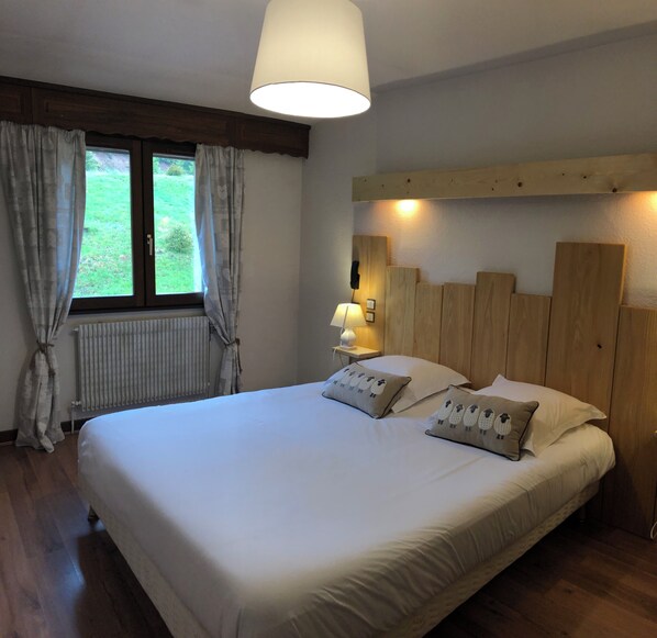 A bedroom with a large bed, wooden headboard, a bedside lamp, and a view of the outdoors through a window with curtains.