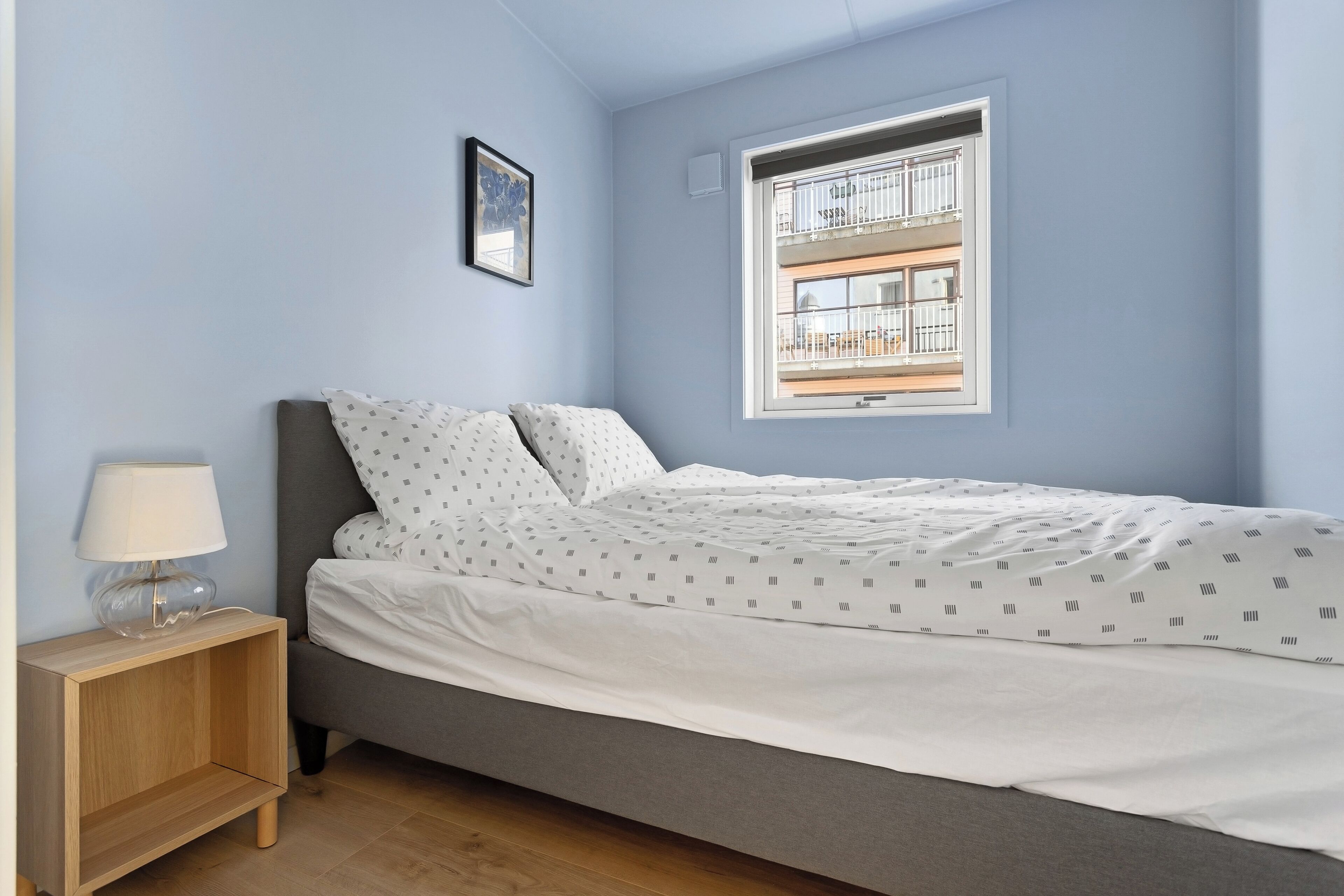 Ein ordentlich bezogenes Bett mit einem gemusterten Überwurf, ein Nachttisch mit Lampe und ein Fenster mit Blick auf einen Balkon.
