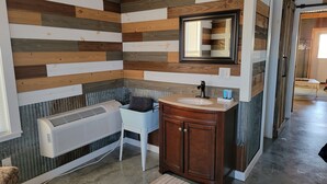 A bathroom with a wooden wall panel design, a white sink, a mirror, and a towel rack. - Foxy's Den cozy grain bin cabin with WiFi & AC in serene Kidder. Quiet & serene. (Kidder)