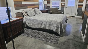 A bedroom with a bed, a nightstand, a lamp, a chair, and a door. - Foxy's Den cozy grain bin cabin with WiFi & AC in serene Kidder. Quiet & serene. (Kidder)
