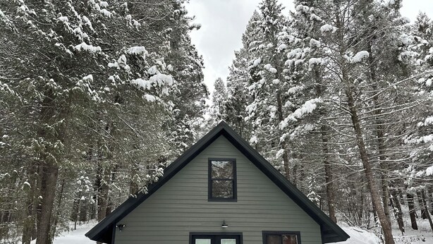 A small, single-story cabin with a stone foundation and a gabled roof, surrounded by snow-covered trees.