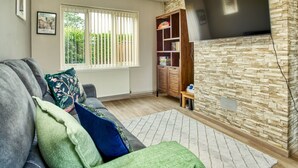 A living room with a grey sofa, a green throw blanket, a white rug, a wooden bookshelf, and a flat-screen TV mounted on a stone wall.