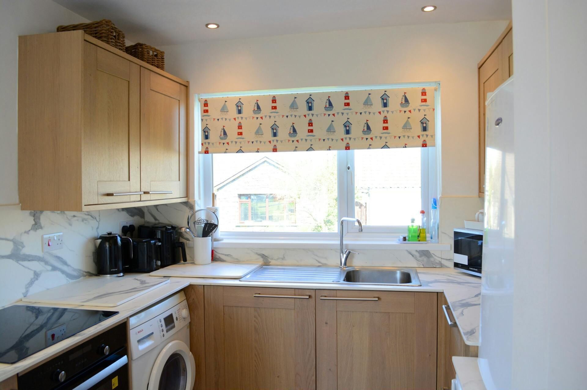A compact kitchen with wooden cabinets, a built-in oven, a microwave, and a window with a nautical-themed curtain.