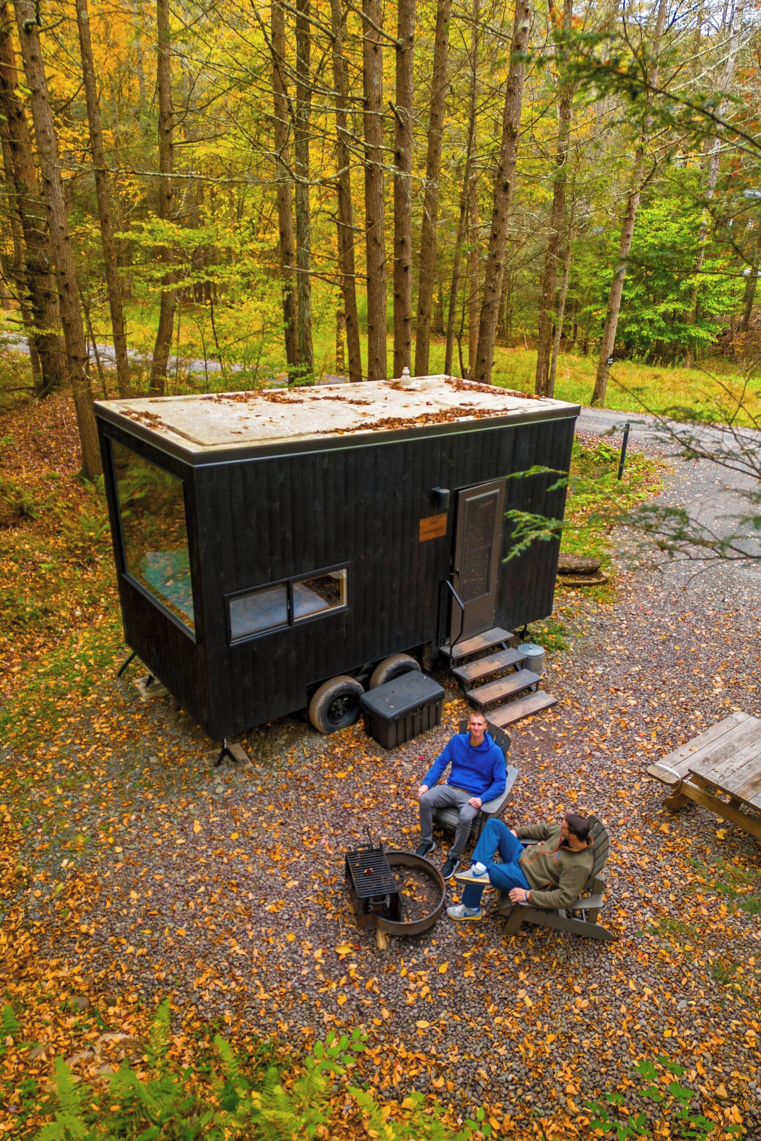 Une petite cabane moderne, dotée d’une grande fenêtre et d’une porte, située dans une zone boisée, le long d’un chemin de gravier.