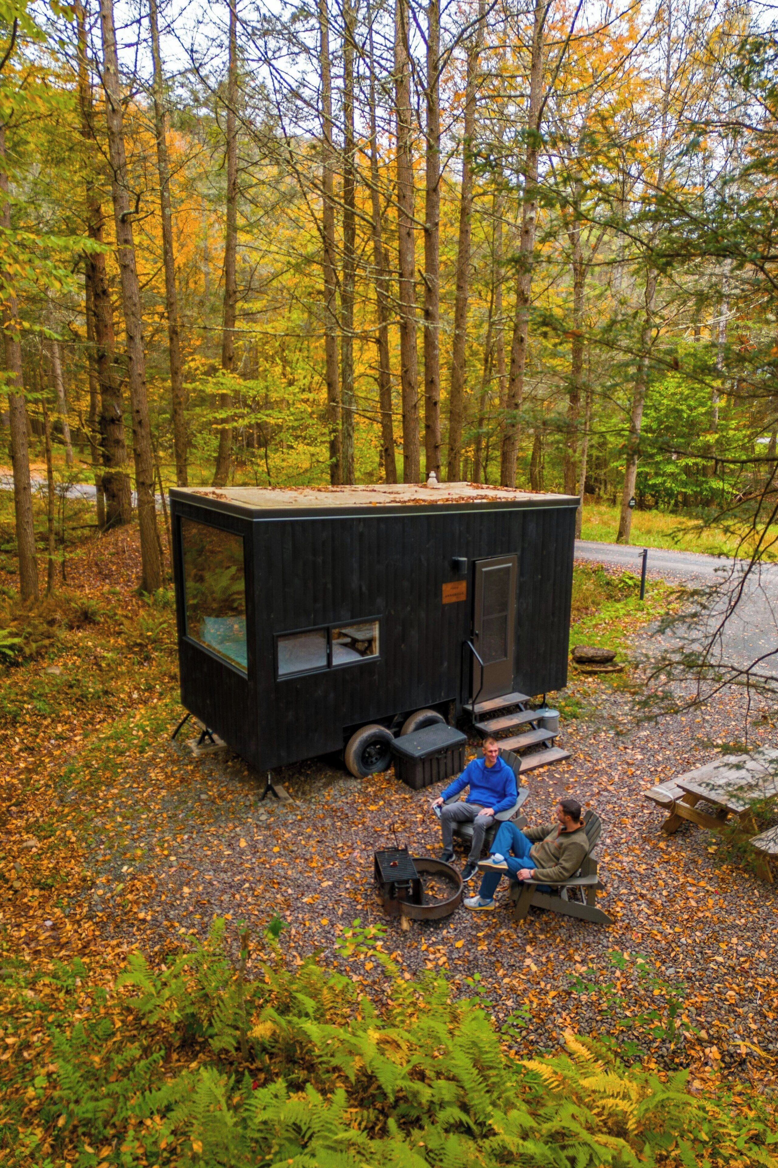 A small cabin with a front window and a door, situated in a forest with trees and fallen leaves.