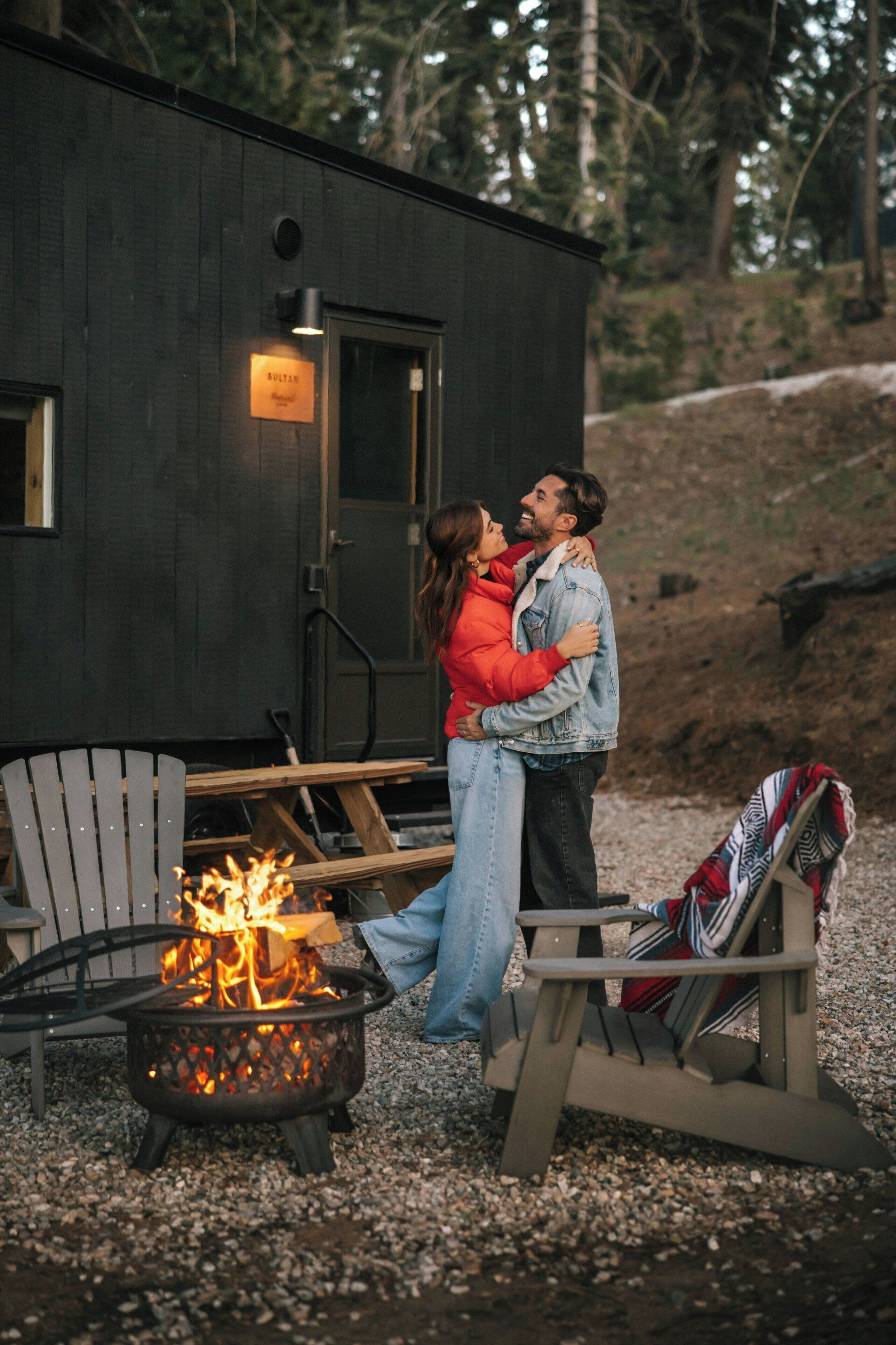 A couple embracing near a fire pit in an outdoor setting with a wooden building in the background.