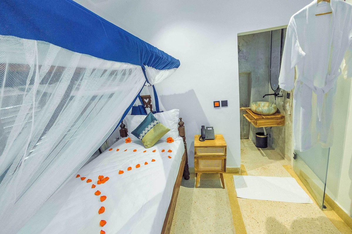 Habitació d'hotel senzilla amb llit, taula de nit petita, telèfon i bany visible a través d'una porta oberta.