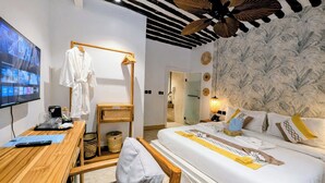 Una habitación de hotel con un escritorio de madera, una cama con almohadones decorativos, un televisor en la pared y un ventilador de techo.