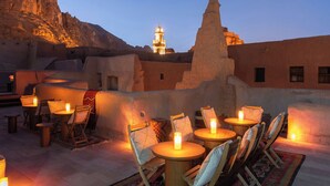 An outdoor seating area with tables and chairs, a mountain backdrop, and a lit tower in the distance. - Dar Tantora The House Hotel (Al-'Ula)