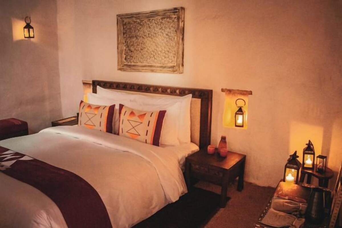 Una cama bien hecha con cabecera, una mesita de noche con lámpara y una lámpara de pared.