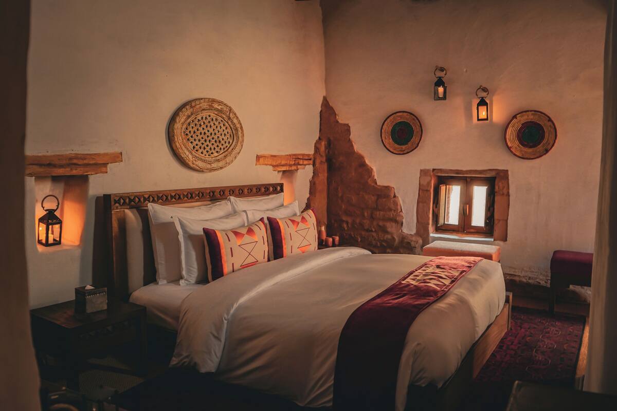 Quarto com uma cama grande, cabeceira de madeira, parede de pedra e duas lanternas fixadas na parede.