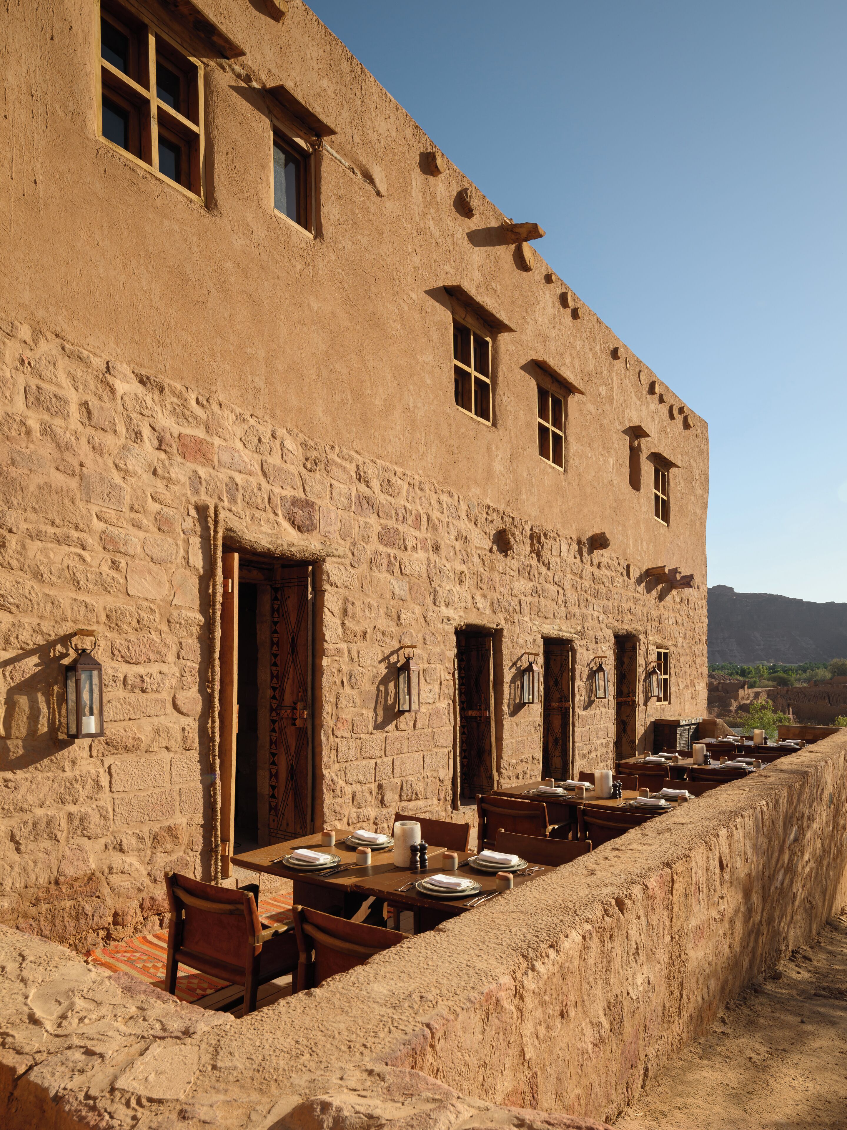 Un edificio con varias ventanas y puertas, mesas y sillas para exterior, y un muro de piedra.