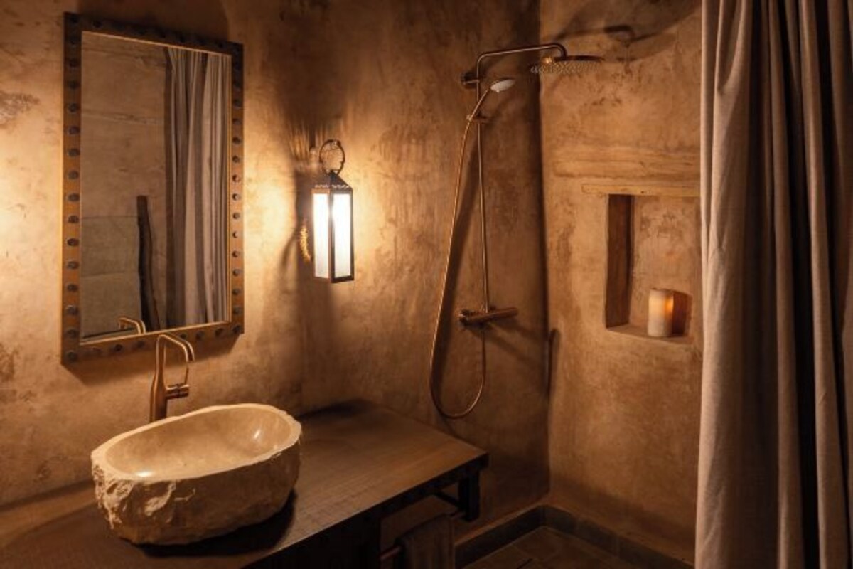 Une salle de bain rustique avec un lavabo en pierre, un miroir, une douchette et un luminaire fixé au mur.