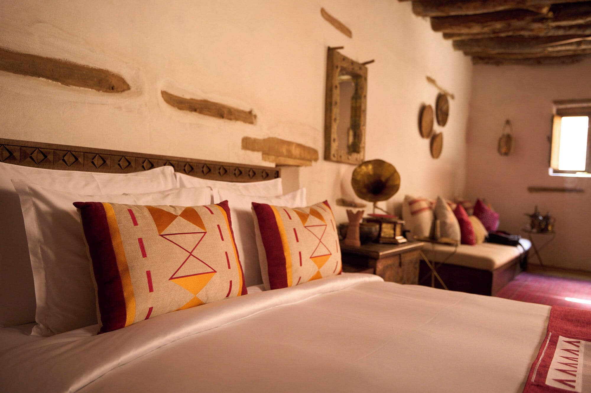 A bed with patterned pillows, a wooden headboard, and a rustic wall with a mirror and decorative items.
