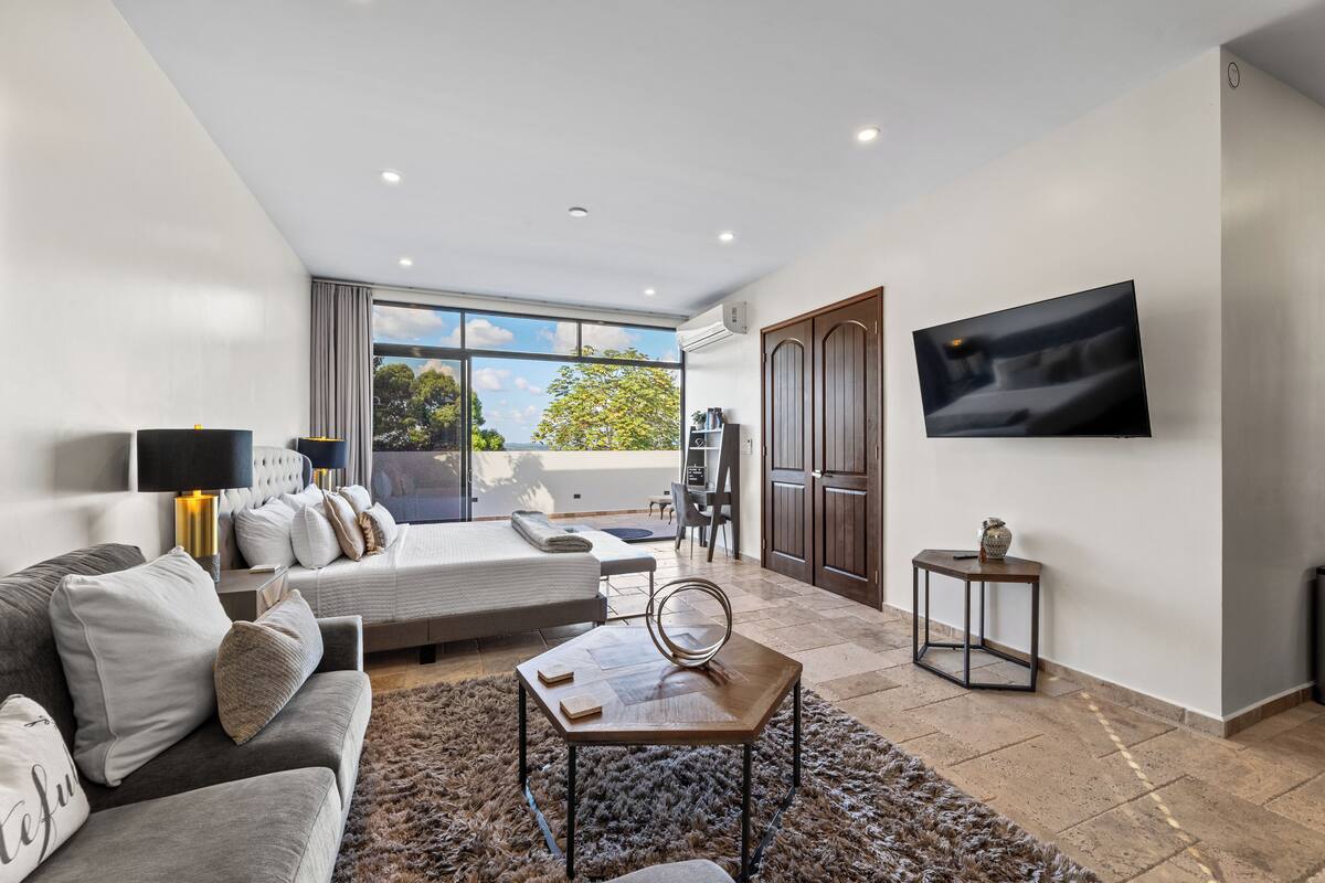 A modern living room with a sofa, a coffee table, a TV, and a large window offering a view of the outdoors.
