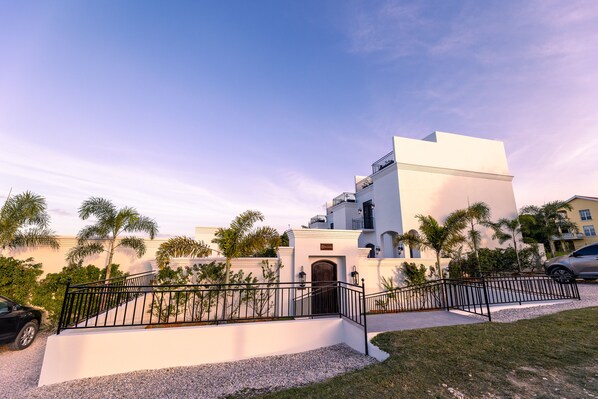 A modern building with a white facade, a gated entrance, and palm trees in the foreground. - Las Haciendas (San Ignacio)