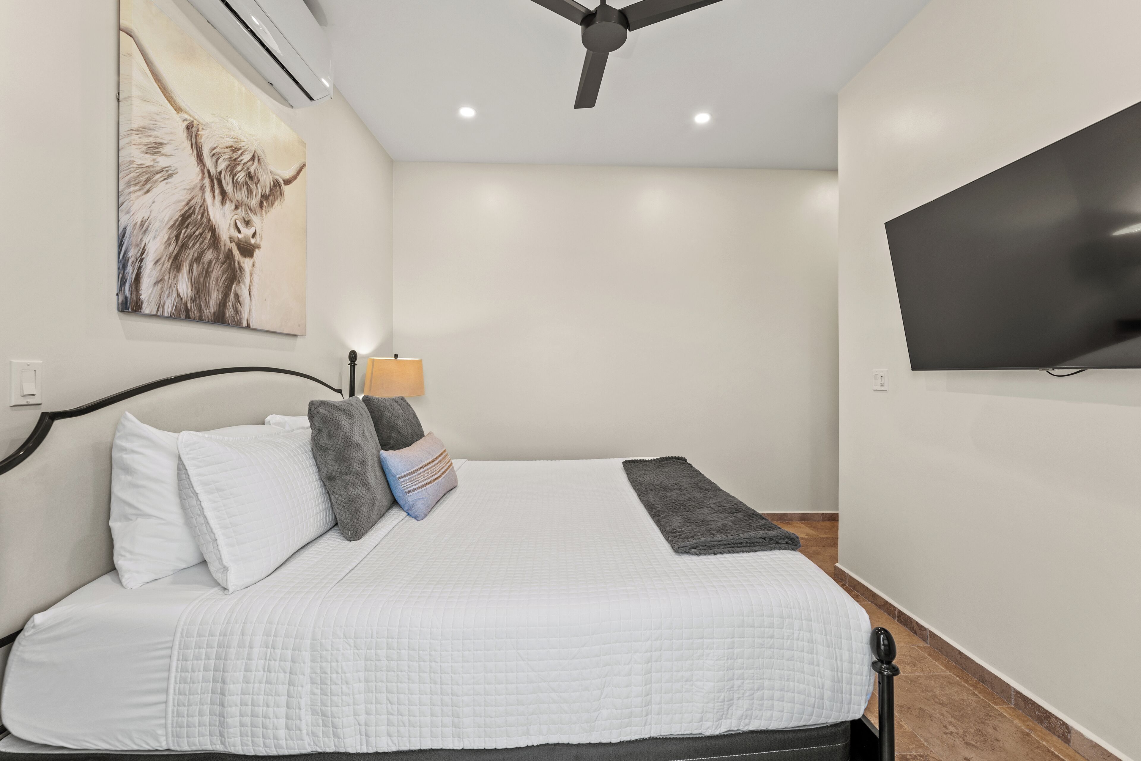 Un dormitorio con cama, almohadas, una lámpara, ventilador de techo, televisor y un cuadro en la pared.