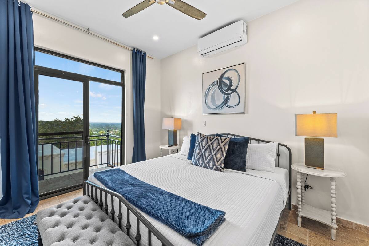 A bedroom with a large bed, two bedside tables, a ceiling fan, and a view of the outdoors.