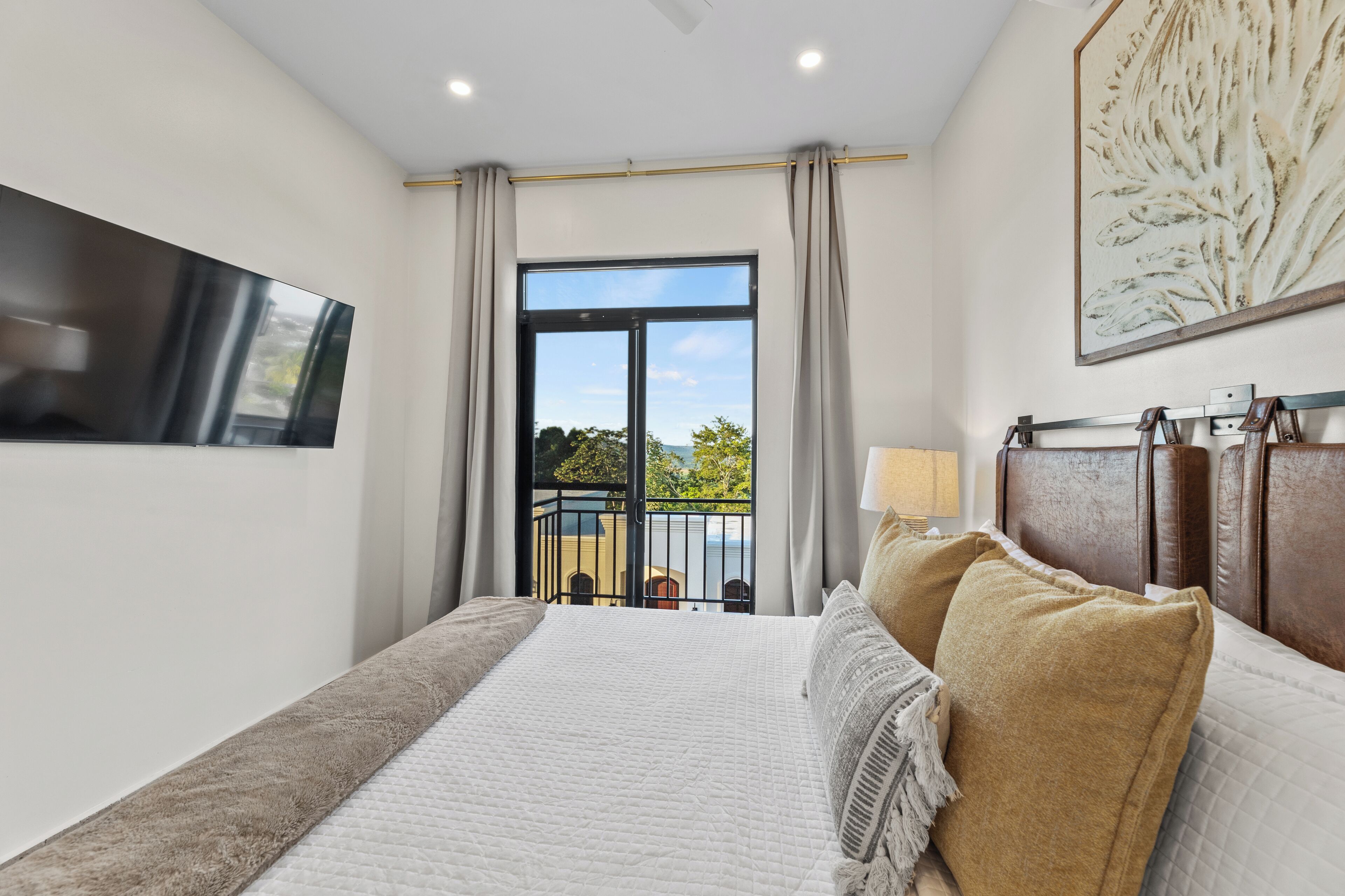 A bedroom with a large bed, a TV mounted on the wall, a balcony with a view of the outdoors, and a painting on the wall.