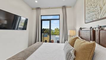 A bedroom with a large bed, a TV mounted on the wall, a balcony with a view of the outdoors, and a painting on the wall.