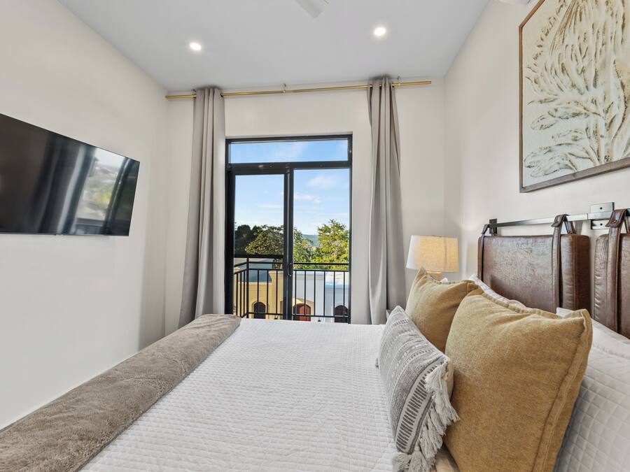 A bedroom with a large bed, a TV mounted on the wall, a balcony with a view of the outdoors, and a painting on the wall.