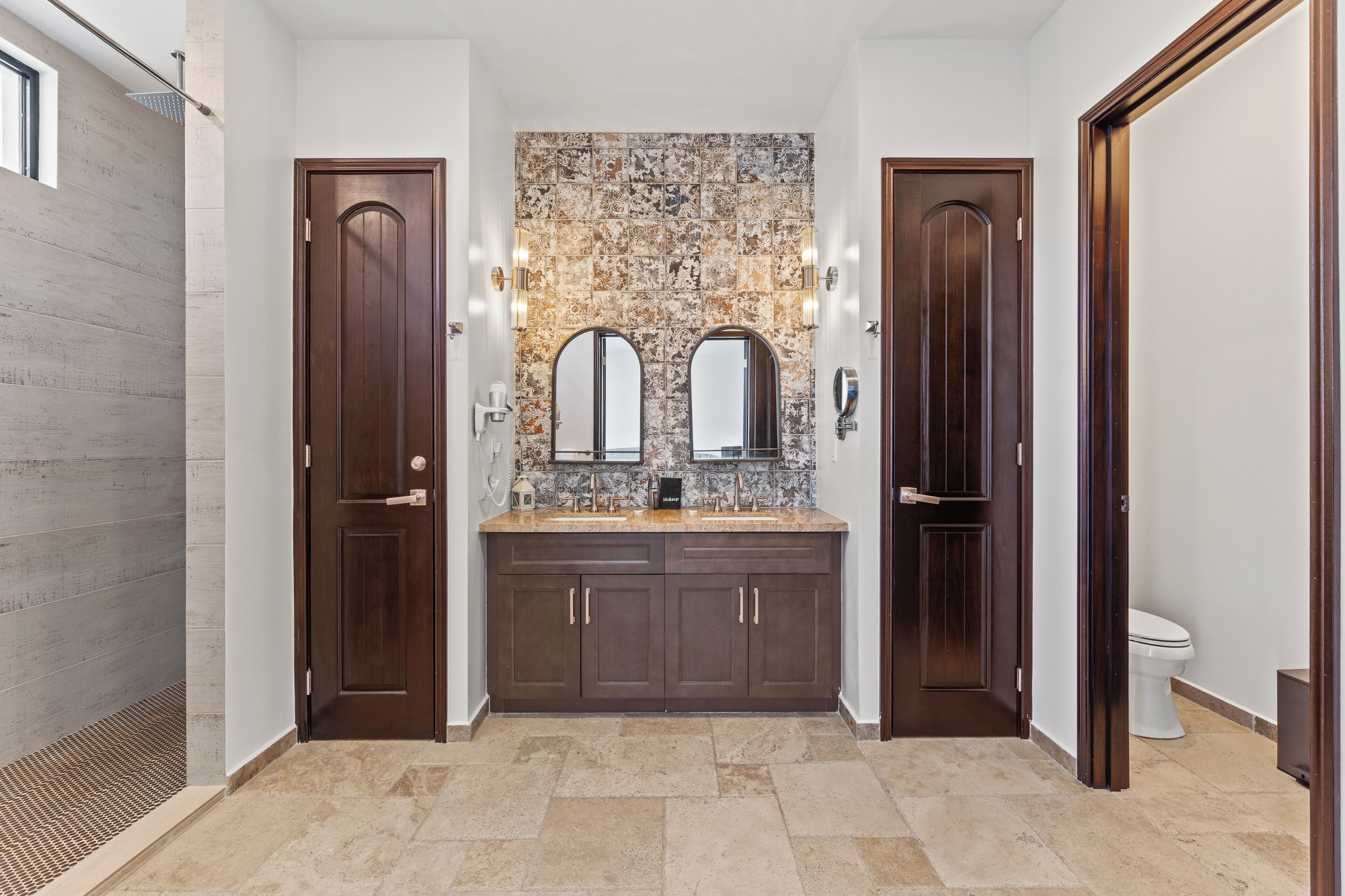 Une salle de bain moderne avec un mur recouvert de carreaux, deux lavabos, des meubles en bois foncé et des toilettes.