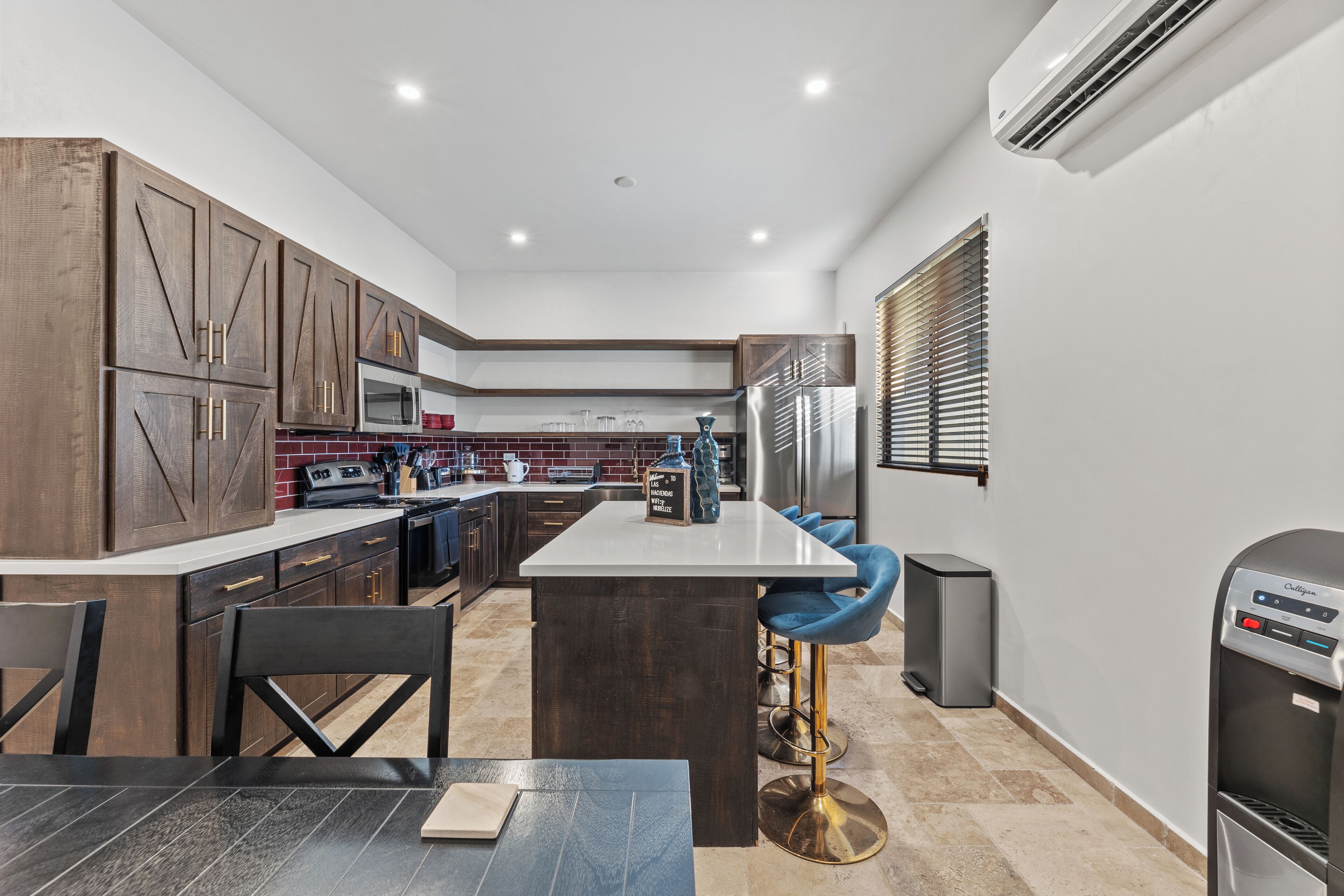 A modern kitchen with dark wood cabinets, a central island with stools, a stainless steel refrigerator, and a water dispenser.