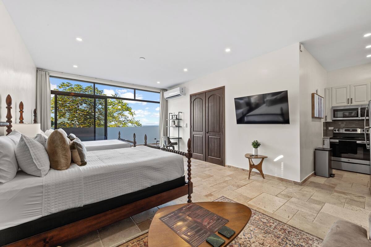 A bedroom with a large bed, a TV mounted on the wall, and a view of the outdoors through a large window.