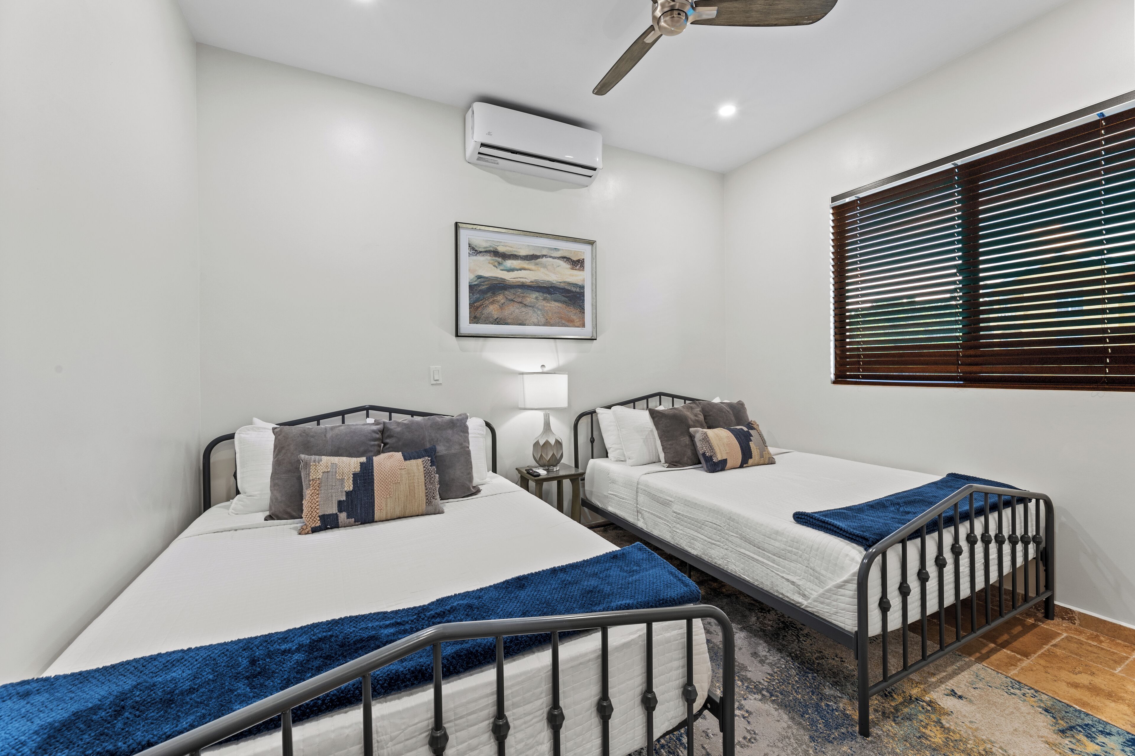 Un dormitorio con dos camas, ventilador de techo, aire acondicionado, un cuadro y una ventana con persianas.