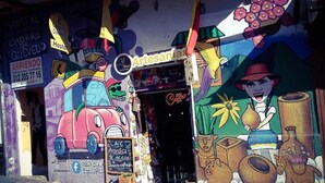 A colorful mural with a red car and a woman holding a pot, a sign for 'Café Artesanal' with a coffee sign, and a 'Hostal' sign with a phone number. - Hostal Chorro De Quevedo Candelaria (Bogotá)