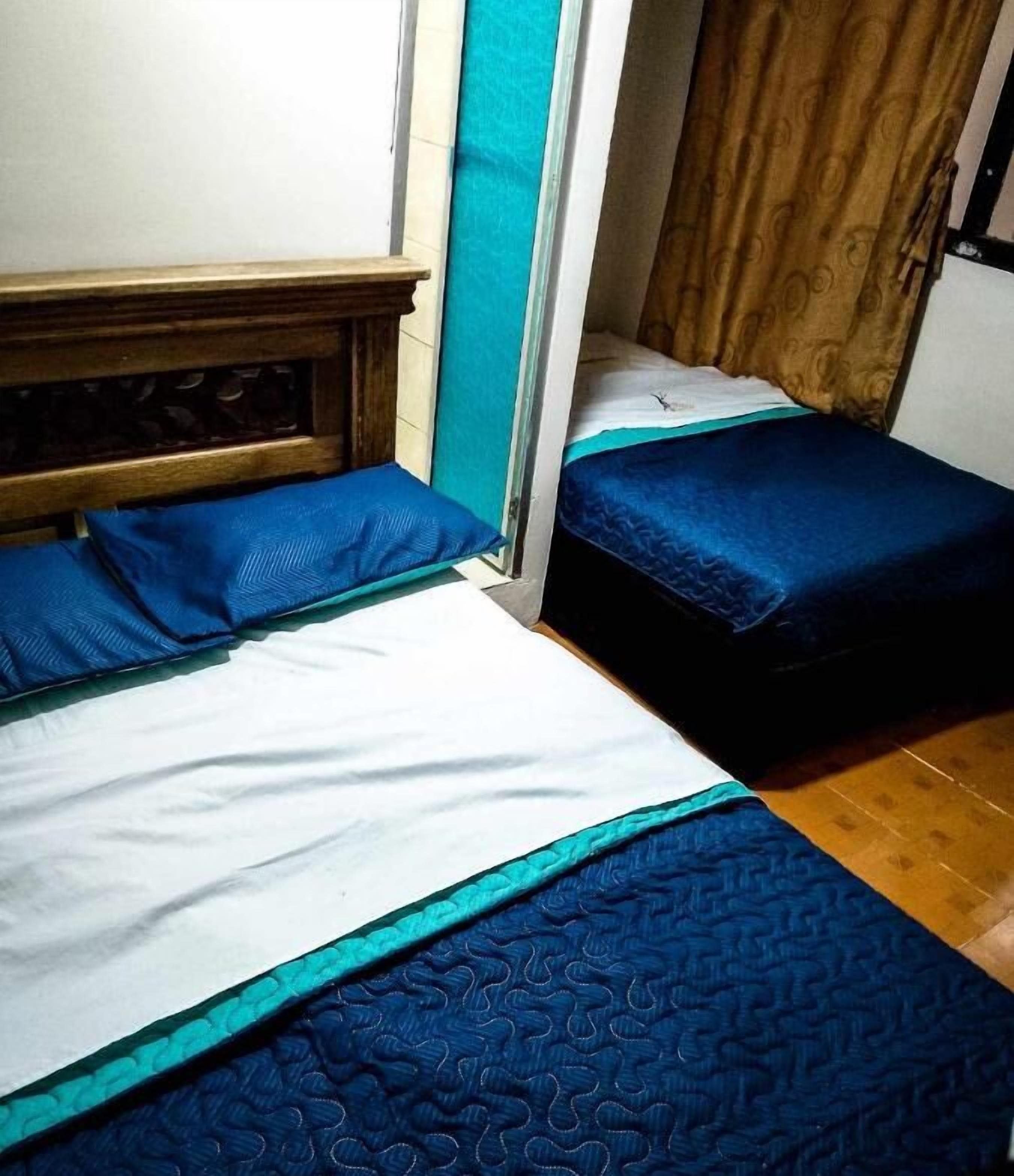 A small, compact room with two beds, each with blue bedding and a wooden headboard. The room has a simple, functional design with a wooden floor and a curtain on one side.