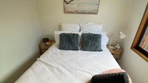 A neatly made bed with white bedding and two dark gray pillows, a pink knitted blanket, and a small bedside table with a lamp and decorative items. - LUXURY 4 BEDROOM KING BED SPA ARCADE WIFI NETFLIX SLEEPS 10 WITH BOAT PARKING (Portland)