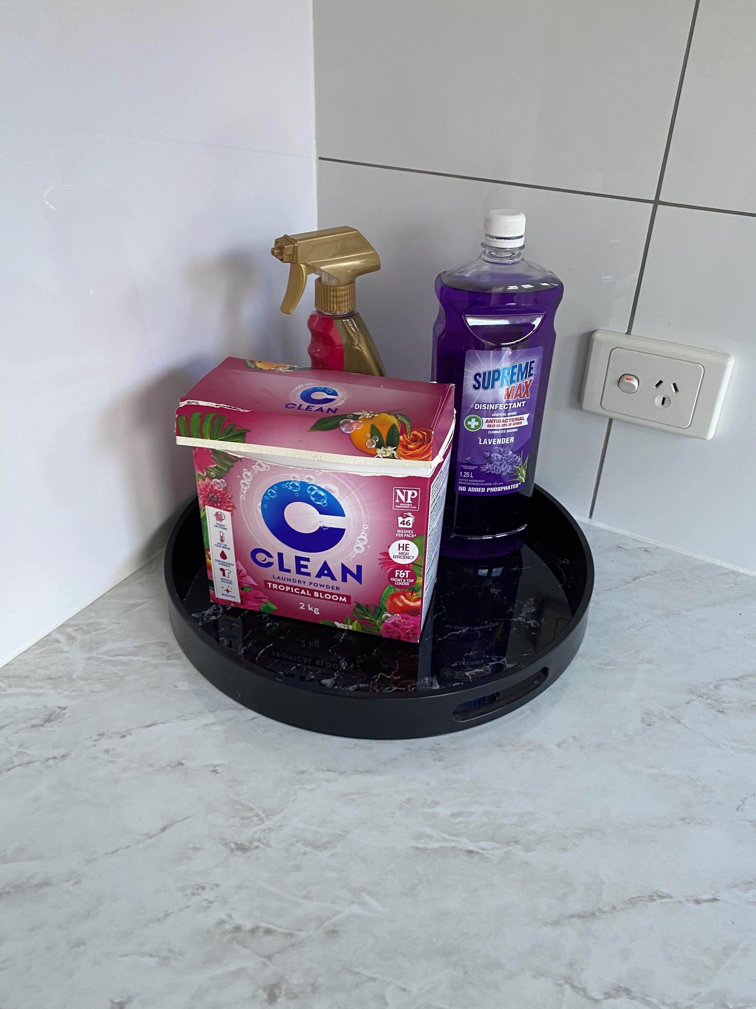 Een zwarte, ronde dienblad op een marmeren aanrecht met een doos 'CLEAN' waspoeder en een fles 'SUPREME MAX' ontsmettingsmiddel.